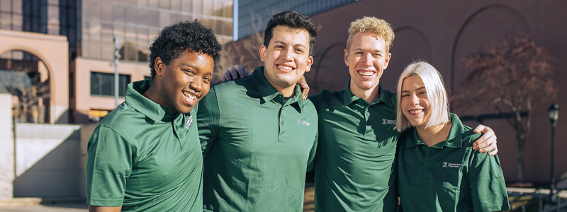 Three male Ensign College students and one female Ensign College student all wearing green Ensign College polo shirts