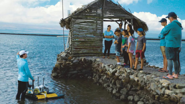 Many people on a dock in the ocean. One person in the water with equipment. 