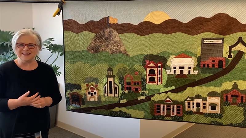 woman standing by a quilt she made that visualizes the history of Ensign College in honor of the college’s new name. 