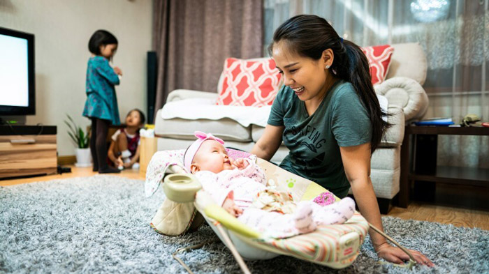 A woman is smiling and looking at her baby while two other children play in the background. 