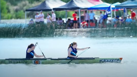 Two young women paddling a large canoe with spectators in background.