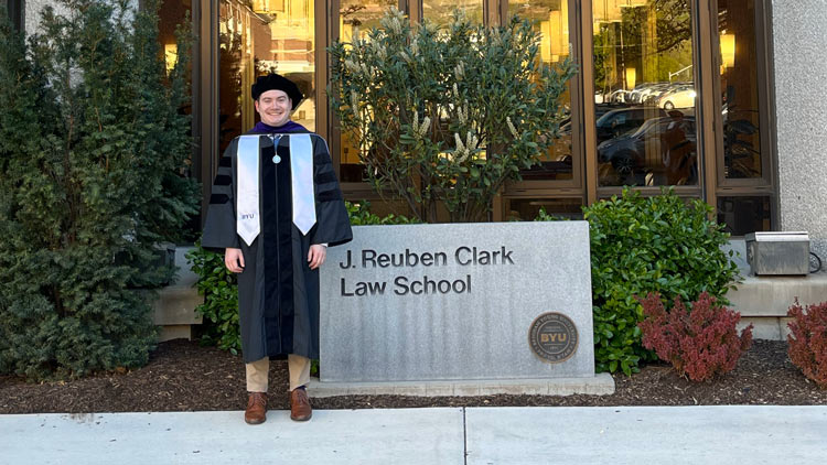 William Emery in his graduation cap and gown standing in front of the J. Reuben Clark Law School building and sign