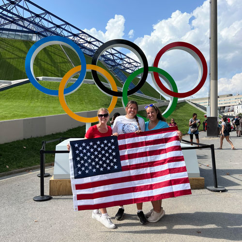 Three students with American flag in front of Olympic rings