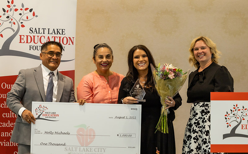 School administrators awarding an over-sized check to a teacher who is holding flowers and a plaque.